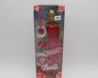 May include: A Barbie doll in a red dress with a heart pattern and a red purse. The doll is packaged in a box with a pink and red heart design. The box says "With Love" and "Barbie".