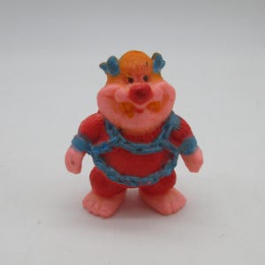 May include: A small, colorful figurine of a cartoon character. The figure has a pink face with a red nose and orange hair. It wears a red outfit with blue accents and a blue chain design across the chest.