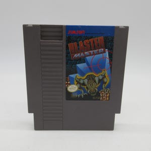 Könnte beinhalten: Eine graue Nintendo Entertainment System (NES) Spielkassette für "Blaster Master". Das Etikett zeigt den Spieltitel in Rot und eine Monstergrafik. Das Sunsoft-Logo befindet sich oben. Die Kassette ist rechteckig.