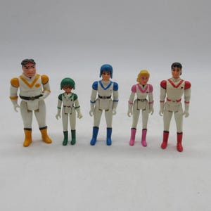 May include: A set of five plastic action figures wearing white uniforms with colored accents. The figures feature distinct color schemes including yellow, green, blue, pink, and red on their helmets, shoulder pads, and boots, standing in a row.
