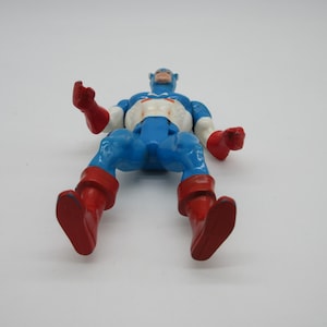 May include: A vintage action figure of Captain America in his classic blue, red, and white costume. The figure is posed with its arms outstretched and legs bent, as if in mid-action. The figure is made of plastic and has a painted finish.