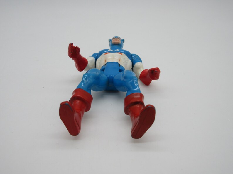 May include: A vintage action figure of Captain America in his classic blue, red, and white costume. The figure is posed with its arms outstretched and legs bent, as if in mid-action. The figure is made of plastic and has a painted finish.