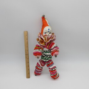 May include: A vintage clown doll with a colourful patchwork costume and a red and orange hat with bells. The doll has a painted face with blue eyes and a smile.