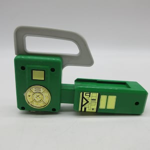Op de afbeelding: Groen en grijs plastic speelgoedgereedschap met een handvat en een wijzerplaat met een ster in het midden. Het gereedschap heeft een gele sticker met een gebouw erop.