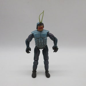 May include: A blue and black action figure with a green antenna, red lips, and a detailed torso. The figure is standing on a white background.