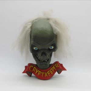 Peut inclure: Une figurine du Gardien de la Crypte à la peau verte, avec des cheveux blancs et des yeux bleus. La figurine porte une bannière rouge avec l'inscription "Crypt Keeper" en lettres jaunes.