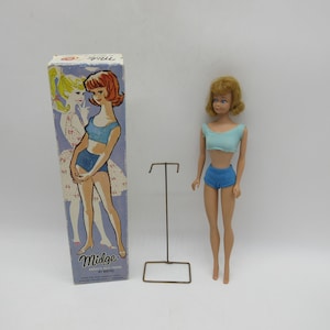 May include: A vintage Midge doll wearing a blue swimsuit and a blue and white striped top. The doll is standing on a metal stand. The doll is packaged in a box with a picture of Midge on the front.