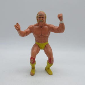 Può includere: Un action figure in plastica di un wrestler muscoloso con capelli biondi, baffi e una fascia bianca sulla testa. Il personaggio indossa slip gialli, stivali gialli con dettagli rossi e polsini bianchi, in posa con le braccia sollevate e flesse.