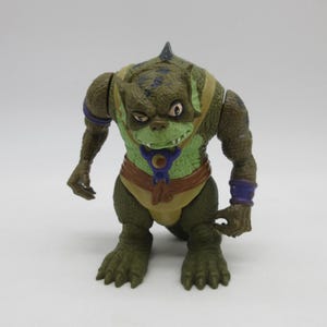 Puede incluir: Una figura de acción verde y marrón de una criatura con apariencia de reptil. La figura tiene una aleta en la cabeza, una cara verde y viste un atuendo amarillo y marrón. Tiene pulseras moradas y un collar azul.