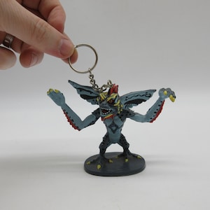 May include: A blue and red Gremlin keychain with yellow eyes and a black base. The Gremlin is standing with its arms outstretched and has a menacing expression.