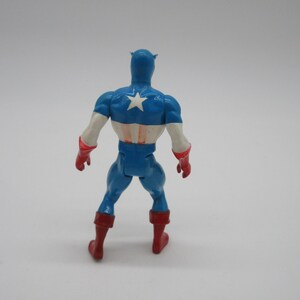 May include: A blue and red plastic action figure of Captain America. The figure is standing with its back to the camera. The figure is wearing a blue and red costume with a white star on the chest. The figure has a red and white helmet.