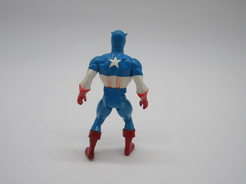 May include: A blue and red plastic action figure of Captain America. The figure is standing with its back to the camera. The figure is wearing a blue and red costume with a white star on the chest. The figure has a red and white helmet.