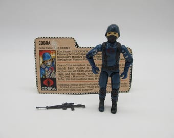 Puede incluir: Una figura de acción vintage de Cobra con uniforme azul oscuro, máscara y casco negros. También hay un pequeño rifle negro y una tarjeta de archivo Cobra. La tarjeta tiene el logotipo de Cobra y texto.