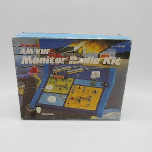 Może przedstawiać: Vintage Science Fair AM-VHF Monitor Radio Kit box. Pudełko posiada niebiesko-żółty projekt obwodu radiowego z napisem "AM-VHF Monitor Radio Kit". Pudełko ma ilustrację strażaka i płonącego budynku.