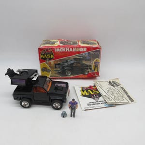 Peut inclure: Un camion jouet M.A.S.K. Jackhammer noir avec un mécanisme de toit transformable, une petite figurine en chemise violette, un casque gris, la boîte d'emballage en carton d'origine avec des graphismes rouges et jaunes, et un manuel d'instructions.