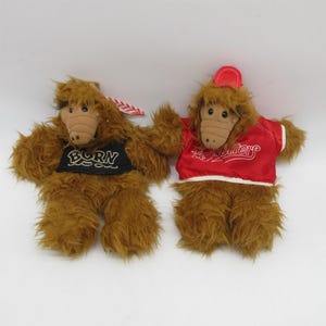 May include: Two plush ALF dolls. One doll wears a black patch with the word "BORN" in white. The other doll wears a red jacket with white trim and the word "ALF" in white script. Both dolls have brown fur.