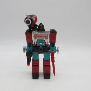 May include: A robot action figure featuring a red, black, and teal colour scheme. The figure has a large cannon mounted on its right shoulder, a clear chest panel, and articulated limbs. It stands in a vertical position with a black head piece.