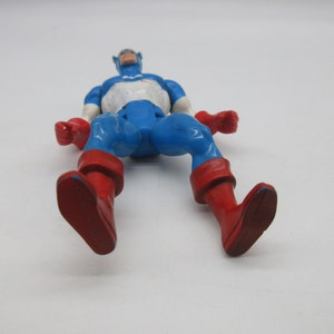 May include: A blue and red plastic action figure of Captain America. The figure is lying on its back with its arms outstretched and legs bent at the knees. The figure has a white star on its chest and a red, white, and blue helmet.