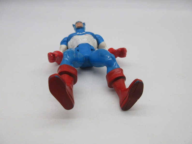 May include: A blue and red plastic action figure of Captain America. The figure is lying on its back with its arms outstretched and legs bent at the knees. The figure has a white star on its chest and a red, white, and blue helmet.