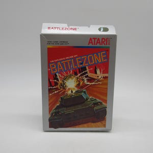 May include: A white Atari game cartridge box with a red and blue graphic of a tank and the text "BATTLEZONE" on the front. The text "THE EXPLOSIVE ARCADE HIT!" is also on the front of the box.