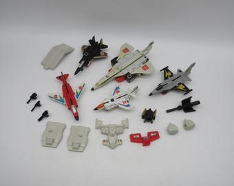 May include: A collection of vintage Transformers toys in their jet vehicle forms. The toys are mostly white and grey with red, black, and yellow accents. The toys include a variety of jet designs, including a fighter jet, a bomber, and a supersonic jet.