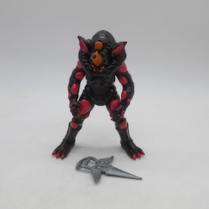 Peut inclure: Figurine d'action de monstre noir et rouge avec une arme grise. La figurine a une grosse tête avec trois yeux et des taches rouges sur son corps. L'arme est une lame argentée.