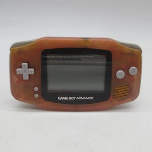 May include: A handheld Game Boy Advance video game console featuring a translucent orange plastic shell. The device includes a central screen, a light gray directional pad on the left, and two light gray A and B buttons on the right side.