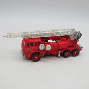 May include: A red toy fire truck featuring a long, white, extendable ladder mounted on top. The vehicle has black wheels, silver-colored accents on the side, and a detailed cab section with a small emblem printed on the front side panel.