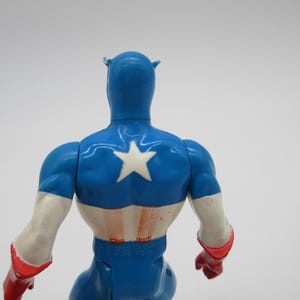 May include: A blue and red plastic action figure of Captain America. The figure is wearing a blue and white costume with a red star on the chest. The figure is facing away from the camera.