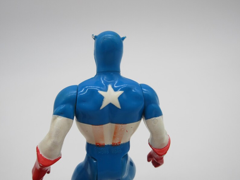 May include: A blue and red plastic action figure of Captain America. The figure is wearing a blue and white costume with a red star on the chest. The figure is facing away from the camera.