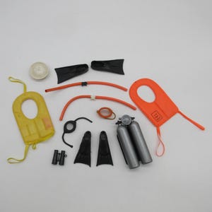 May include: A collection of miniature scuba diving accessories including a yellow life vest, an orange life vest with the text AIR SEA RESCUE, two pairs of black swim fins, a silver double oxygen tank, black binoculars, two orange tubes, and a white circular disc.