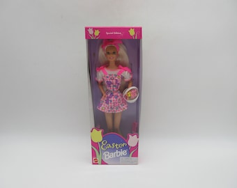 May include: A pink and white Easter Barbie doll in a pink dress with a floral pattern and a basket of Easter eggs. The doll is in a clear plastic box with a pink and white background. The box has the words "Easter Barbie" on it.
