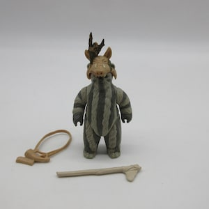 Op de afbeelding: Een grijs en bruin action figure van een wezen met een gehoornd hoofd en een gestreept lichaam. De figuur houdt een stokachtig wapen vast.