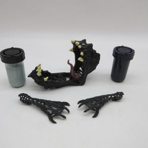 May include: A black plastic monster figure with a large open mouth, two small arms, and two small legs. The figure is broken into five pieces. There are two small black plastic containers with lids.