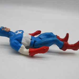 May include: A vintage action figure of Captain America lying on its back. The figure is made of plastic and is painted in the classic red, white, and blue colors. The figure has a star on its chest and a red, white, and blue helmet.