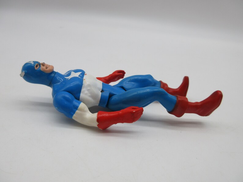 May include: A vintage action figure of Captain America lying on its back. The figure is made of plastic and is painted in the classic red, white, and blue colors. The figure has a star on its chest and a red, white, and blue helmet.