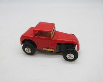 Pode incluir: Um pequeno carro de brinquedo vermelho com teto rígido e pneus pretos. O carro tem um design clássico com uma faixa amarela na lateral. As rodas são pretas com aros prateados. É uma réplica em miniatura.