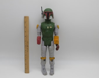 Puede incluir: Una figura de acción de Boba Fett de Star Wars verde y gris con una pieza de pecho roja y amarilla. La figura está de pie sobre una superficie blanca.
