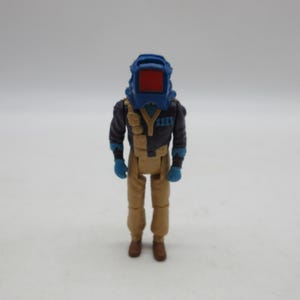 May include: A blue and brown action figure with a blue helmet and a red visor. The figure is wearing a brown jumpsuit and has a black vest with a silver buckle.