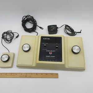 Könnte beinhalten: Eine Vintage-Videospielkonsole von Gloritone mit zwei Controllern, einem Netzteil und zwei Kabeln. Die Konsole ist beige mit schwarzen Knöpfen und einem schwarzen Bildschirm. Die Konsole trägt den Text "Gloritone" und "Game Select".