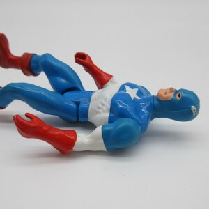 May include: A blue and red plastic action figure of Captain America lying on its back. The figure is wearing a blue and white costume with a red star on the chest. The figure has a red and white mask.