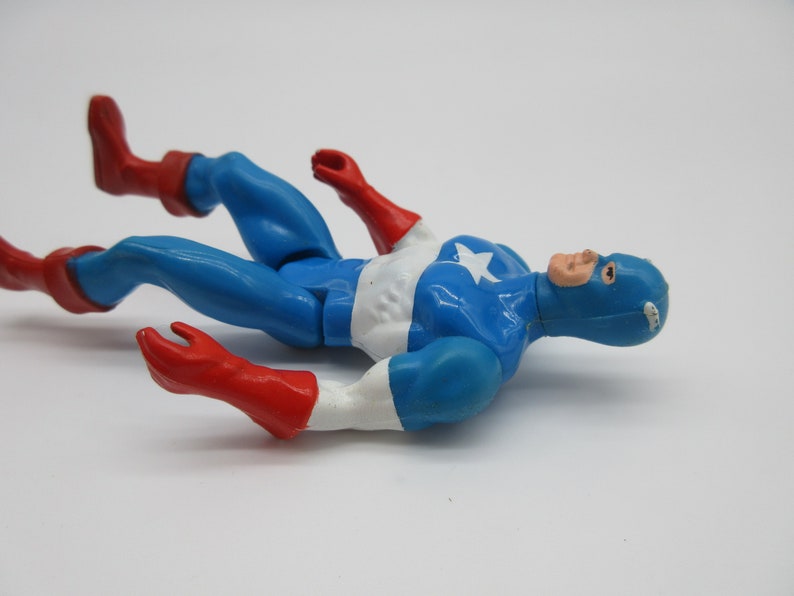May include: A blue and red plastic action figure of Captain America lying on its back. The figure is wearing a blue and white costume with a red star on the chest. The figure has a red and white mask.