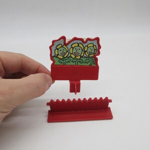 May include: A red plastic toy with a flower design. The top piece features three smiling yellow flowers with green leaves. The bottom piece is a red base with a saw-tooth edge. The toy is designed to stand upright.