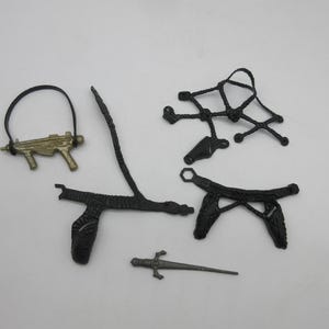 May include: A set of black plastic toy accessories for action figures, including a gun, a sword, and two belts with holsters.