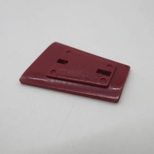May include: A maroon plastic rectangular object with a smaller, raised section on top. The raised section has two rectangular cutouts and four small holes. The object is likely a component or part of a larger item.