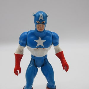 May include: A vintage action figure of Captain America, wearing a blue, white, and red costume with a red star on his chest. The figure has a red and white mask with the letter "A" on the front.