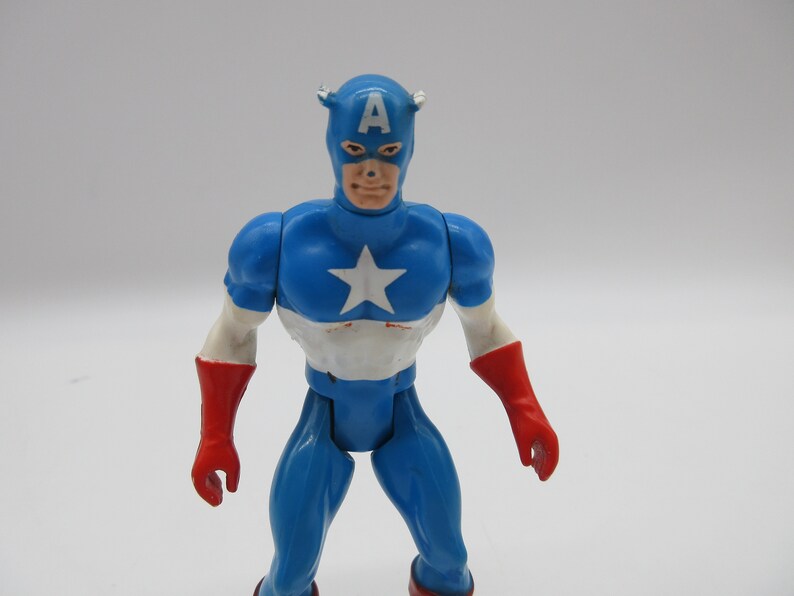May include: A vintage action figure of Captain America, wearing a blue, white, and red costume with a red star on his chest. The figure has a red and white mask with the letter "A" on the front.