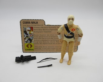 Puede incluir: Figura de acción de Storm Shadow con accesorios, incluyendo un arco, espada y arma. La figura es de color beige y blanco, con una faja negra. Una tarjeta de Cobra Ninja está en el fondo.