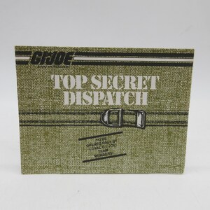 May include: A rectangular, olive green box with the text "G.I. JOE TOP SECRET DISPATCH" in white. The box has black horizontal lines and a silver buckle graphic. The bottom of the box reads "TO BE OPENED ONLY BY LOYAL G.I. JOE TEAM MEMBERS."