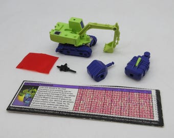 May include: A green and blue toy digger with a red square, two blue accessories, a black accessory, and a card with text. The digger has a movable arm and a scoop. The card has text that reads "CONSTRUCTION MINING & SALVAGE".